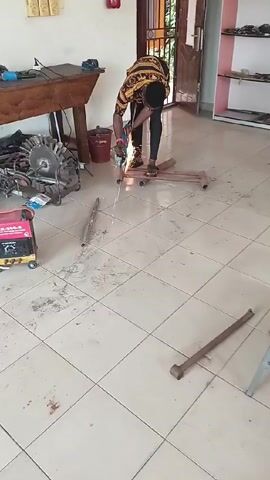 A man wearing a patterned shirt, dark pants, and sandals is standing indoors on a tiled floor, using an angle grinder to cut a piece of metal pipe. Sparks are flying as he works. Several pieces of metal pipe are scattered around him on the floor, along with a small generator. There is a wooden table with tools on it in the background, as well as a red bucket and a metal shelf unit. The room appears to be a workshop or garage area.