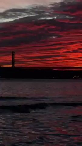 The video shows a seascape at sunset. The sky is a fiery red, streaked with dark clouds. A silhouetted bridge is visible on the horizon. The water is dark and choppy, with small waves.