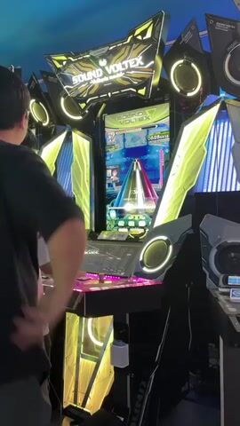 A person is playing a music arcade game called Sound Voltex. The game is a rhythm game where players press buttons and turn knobs to match on-screen cues. The machine is brightly lit with colorful LEDs. The person is focused on the game, with their hands on the controls. The environment appears to be an arcade or entertainment venue, with other similar machines visible in the background.