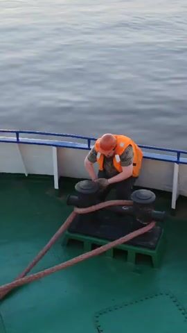 A man wearing a hard hat, a camouflage t-shirt, and an orange life jacket is on a boat, securing a thick rope around a bollard. The boat deck is green, and the railing is white and blue. The sea stretches out behind him.