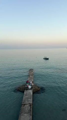This is a vertical video of a serene seascape at sunset. The primary subjects are a small group of people gathered on a concrete pier that extends into calm, turquoise water. They appear to be enjoying a picnic or relaxing, with some seated and others standing. The pier is weathered and leads to a small, rocky outcrop where the group is concentrated.

In the distance, a small white boat with a dark hull is anchored or drifting in the tranquil sea. The water itself is clear, allowing a view of the seabed in shallower areas. The sky displays a gradient of soft colors, transitioning from a pale blue near the horizon to a light pinkish-orange higher up, indicative of either sunrise or sunset. There are a few buoys visible in the water, marking the expanse of the sea. The overall mood is peaceful and tranquil.

The setting appears to be a coastal area, given the pier, the sea, and the distant hint of land on the far right of the frame, suggesting a shoreline or island. The time of day is clearly either dawn or dusk due to the sky's coloration and the soft light. The weather appears to be calm, with no visible wind or rough seas. No text is visible in the media.