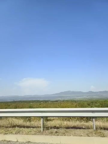 The video shows a view from a moving vehicle looking out over a landscape under a clear blue sky. In the foreground, there is a white guardrail along the side of what appears to be a road. Beyond the guardrail, there is dry grass and some sparse vegetation. In the middle ground, there is a vast expanse of green vegetation, possibly fields or a natural landscape. In the background, there are distant hills or mountains. A single, large white cloud is visible in the sky above the landscape. The overall scene depicts a bright, sunny day in a rural or semi-rural setting.