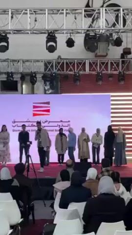 Certainly! Here's a description of the scene in the video:

The video shows a group of people on a stage at what appears to be the Damascus International Book Fair. The stage has a large screen in the background with the fair's logo and name in Arabic and English.

A man in a checkered shirt stands center stage speaking into a microphone. To his left, there is a young woman in a floral dress and a young boy in a sweater. To his right, there are six women, some wearing headscarves. In front of the stage, there is an audience seated in rows of white chairs. A camera on a tripod is visible to the left of the speaker. The environment seems to be indoors under a metal structure with lighting fixtures.