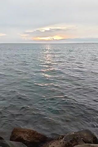 The video shows a view of the ocean at sunset from a rocky shoreline. The water is calm with gentle ripples, reflecting the sunlight. The horizon is visible with a sliver of bright light peeking through the clouds. The sky is overcast.  No people are visible.