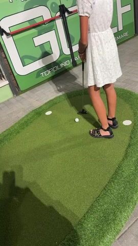 The video shows a group of people playing mini golf. In the first few seconds, a young person in a white dress and sandals is shown putting a golf ball. The background shows a green sign with "GOLF" written on it, and the words "TOPOLINO BARI MARE". In the subsequent seconds, the camera pans to show other people playing mini golf, including a child in shorts and sandals, and adults. The mini golf course is made of green artificial turf with white holes. There are various obstacles on the course, including a large white golf ball-shaped structure. The ground surrounding the mini golf course is paved. The lighting suggests it is either evening or indoors.