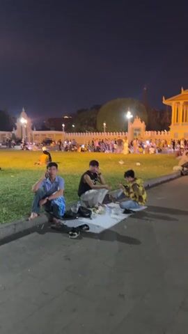 The video shows a nighttime scene in what appears to be a public square in front of a building illuminated with bright yellow lights. There are many people gathered on a large grassy area. Some are walking, while others are sitting on the grass on mats or blankets. Three young men are seated on a piece of cardboard on the edge of the walkway, eating. Several water bottles and bags are near them. The overall atmosphere appears relaxed and social, with the brightly lit building serving as a backdrop for the gathering.