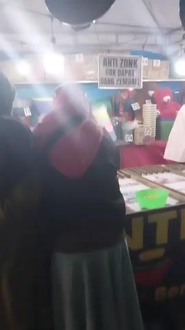 The video shows a game stall at what appears to be a fair or market. In the foreground, two people are visible from behind, facing the stall. The person on the left is wearing a red hijab and a black jacket, and the person on the right is wearing a light-colored hijab and a light-colored top. The stall itself is set up with a table displaying a game, possibly a ring toss or a similar game of chance, with green bowls and orange balls. Behind the table, a woman wearing a hijab and a light-colored shirt is attending the stall and interacting with the customers. There are shelves behind her stocked with various items, including what appear to be plastic containers and stuffed toys. A sign on the stall reads "ANTI ZONK GAK DAPAT UANG KEMBALI," which translates to "Anti-zonk, no money back," suggesting a prize game. The lighting is dim, typical of an indoor or evening market setting.