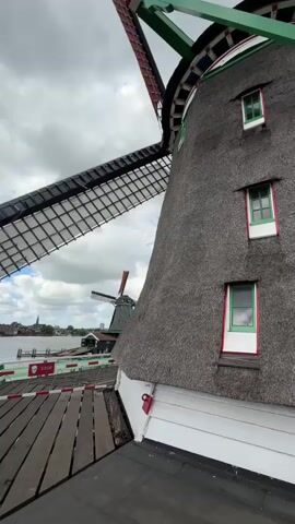 The video displays a close-up of a traditional Dutch windmill with a thatched roof and four small windows. The windmill's sails are partially visible, with one sail in motion, indicating it is operational. Another windmill, smaller and in the distance, is also visible, along with a body of water and a cloudy sky. The foreground shows a wooden platform or walkway, part of the windmill's structure, with a red and white striped railing and a red and white sign that says "STOP". The camera is positioned at an angle, looking upwards at the windmill, emphasizing its scale. The lighting suggests it is daytime.