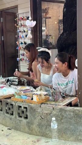 Three young Asian women are gathered around a table, engaged in a craft activity, likely painting or decorating small objects. They are seated outdoors at what appears to be a casual workspace, possibly a workshop or a market stall. The table is cluttered with art supplies, including paint palettes, brushes, containers of paint, and various small decorative items in a tray. One woman, on the left, wears a brown top and is focused on her work. The woman in the center, in a white lace top, is holding a small yellow object, seemingly painting it. The woman on the right, wearing a white t-shirt, is smiling and also holding a small object. The background reveals a rustic building with wooden walls and a display of colorful hair accessories hanging from a rack. A large tree trunk is visible to the right, and a bottle of water sits on a concrete barrier in the foreground. The lighting suggests it is daytime, possibly late morning or afternoon. The overall mood appears relaxed and cheerful. The white t-shirt of the woman on the right has text that reads "weekend". The location context is Ban Sai Kham, Thailand.
