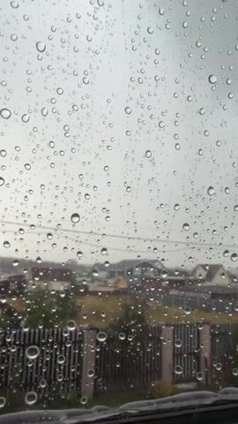 A slow-motion video captures raindrops on a window pane. The raindrops are various sizes and shapes, some appearing as clear spheres, others as elongated streaks. The background is blurred but depicts a residential area with houses and a fence. The sky appears overcast and gray, consistent with rainy weather. The video's focus is on the visual effect of the rain on the glass.