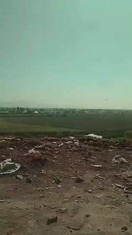 The video shows a scene of a landfill or waste area. There is a person sitting amidst the debris. In the background, there is a view of a distant town or settlement surrounded by green fields. The sky is clear and light. The foreground is dominated by dirt, stones, and scattered waste materials.