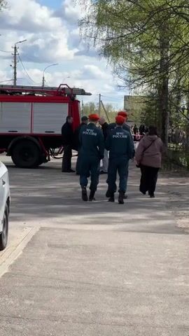 The video shows a fire truck parked on a paved road. Several individuals, including those in dark blue uniforms with "МЧС РОССИИ" (presumably meaning Russian Ministry of Emergency Situations) printed on the back and orange berets, are standing or walking near the truck. Some civilians are also present. The scene appears to be outdoors, with trees and buildings visible in the background under a cloudy sky.