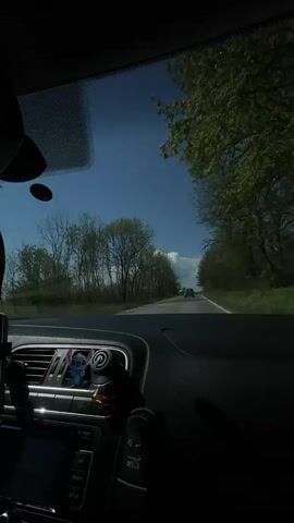 The video shows a drive down a country road. The view is from inside a car, looking out the windshield. The dashboard is visible, and there’s a small Stitch figurine on the center console. The road is narrow, with trees on both sides. The sky is blue with some clouds. Another car is visible in the distance, driving in the same direction. The music is playing.