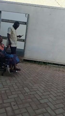 A woman in a wheelchair is being pushed along a brick pathway by a man in blue scrubs. Another man walks alongside them, looking at his phone. The woman is wearing a dark, patterned dress and a headscarf. The man in scrubs is wearing blue scrubs and dark shoes. The other man is wearing a light-colored shirt and dark pants. In the background is a white building with a light-colored roof. A car is parked near the building. The scene appears to be outdoors on a sunny day.