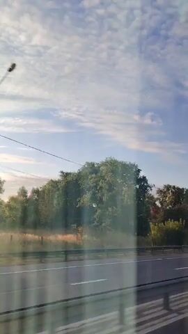 The video shows a view from a moving vehicle looking out the window at a roadside landscape. The road ahead is visible, with a metal guardrail in the foreground. On the right side of the road, there are trees and some buildings. The sky is filled with white, puffy clouds. In one frame, a truck is seen passing by on the road. The overall scene depicts a daytime drive through a semi-rural or suburban area.