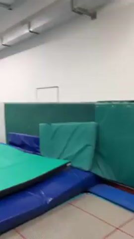 A shirtless young man wearing red shorts does a back flip off a set of green and blue gymnastic crash pads. He lands on another set of crash pads, then stands up, spreading his arms. The scene takes place indoors in a room with white walls, a long white ceiling light fixture, and other gymnastic equipment visible in the background.