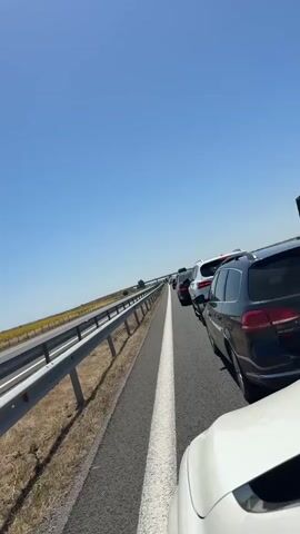 The video shows a line of cars on a highway. The sky is clear and blue. There is a guardrail on the left side of the road and a field of yellow flowers beyond it. The cars are stopped, and it appears to be a traffic jam. The video is shot from the perspective of someone in a car, looking forward.
