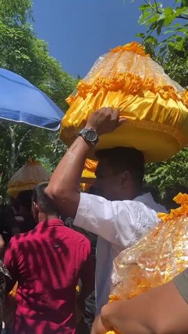 A group of people are seen carrying ornate, bell-shaped objects covered in yellow fabric with ruffled edges. The objects appear to be held atop their heads. The foremost person, wearing a white shirt and a watch, is seen from the side, holding one of these objects steady. Another person in a red shirt is visible in the foreground, also facing away from the camera. In the background, trees and a blue umbrella suggest an outdoor setting under a clear sky. The lighting indicates it is daytime.