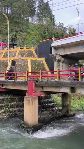 The video shows a bridge over a river. A motorcycle is driving across the bridge. The bridge is supported by concrete pillars. The river has rapids. There is a hillside in the background with trees and vegetation. There is also a building with a yellow and black facade. The sky is overcast.