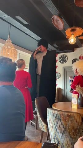 A man wearing a black cape and a large fur hat stands on a raised platform, holding a microphone to his mouth. He is singing to an audience. To his left, a woman in a red dress faces him. To his right, another person in traditional attire with a large white hat is also visible. The background shows a restaurant interior with decorative lamps and wall hangings. The video appears to be a live performance.