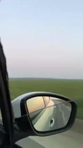 The video shows a view from the passenger seat of a car moving along a road. The side mirror of the white car is visible, reflecting a warm-toned light, possibly from the setting sun. Outside the car, there's a flat, green field stretching out to the horizon under a pale, blue-gray sky. A tall, white structure appears briefly in the distance. The car seems to be driving on a fairly smooth road.