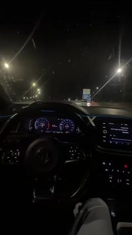 The video shows the interior of a car at night. The driver's perspective is captured, with a steering wheel, dashboard, and infotainment screen visible. The dashboard displays speed and RPMs. The infotainment screen shows song information. The car appears to be moving on a road with streetlights visible outside. The mood is dark and atmospheric.