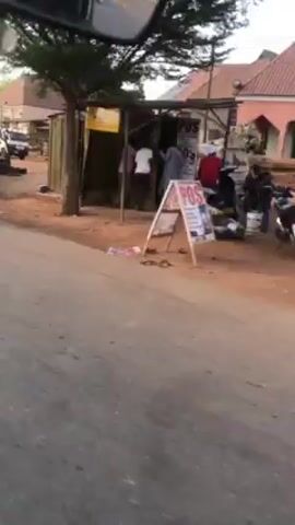 The video shows a car driving down a street in a rural area. There are people walking along the side of the road, and buildings and vegetation in the background. The road is paved, but it is dusty and has some potholes. The car passes several parked vehicles, some of which appear to be in disrepair. There is also a sign advertising a business.
