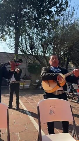 A mariachi band is playing outside on a tiled patio. One member sings and plays a guitar. Others play trumpets and a violin. People sit at tables nearby, listening to the music. There are trees in the background, a brick wall, and a blue sky. The camera moves around the scene.