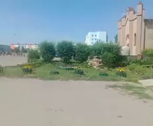 The video captures an outdoor scene with a clear blue sky. In the foreground, there is a sandy or dirt-like ground area. Beyond this, a patch of greenery with bushes and yellow flowers is visible, bordered by what appears to be old tires repurposed as planters. In the mid-ground, a group of people, seemingly in formation and possibly military or cadets, are spread across a paved area. They are standing in rows. To the left, a building with multiple windows and some cars parked in front of it is present. Further back, there are several residential buildings with colorful facades. On the right side of the frame, a light-colored building with a distinctive architectural style, possibly a church or a unique public building, stands out. A flag is visible on a pole in the far left background. The lighting suggests it is daytime, likely sunny.