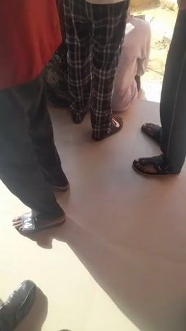Several people stand in a line outside of a building. Most are wearing some type of long-sleeved shirt or jacket and pants. Many are wearing sandals or flip-flops. The person in the center of the frame is wearing a white t-shirt with the words "GIVE ELECTIONS & RELIGIOUS KILLINGS A GOOD KNOCK DON'T KILL OR SUPPORT KILLINGS" printed on the back. The video shows the group moving along in the line and the building to the left and outside area in the background.