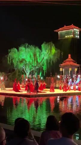 The video shows a nighttime performance on a stage in the middle of a body of water. The stage is lit in red and green, and the reflection of the lights and stage is visible in the water. There are several performers on the stage dressed in red and blue traditional clothing. They are moving around the stage, and some are carrying props like fans. Behind the stage, there is a large green tree and a traditional Chinese-style building with a red roof. Above the building, there is a small floating structure with lights. In the foreground, there are several people sitting and watching the performance. The audience is blurred and their faces are not clearly visible.