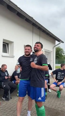 Two men, one holding a bottle and wearing a black t-shirt and blue shorts, the other in a similar outfit, are embracing and cheering outdoors. They are standing on a paved area next to a white building. Other men are visible in the background, some seated, some standing, also wearing similar attire. The sky is overcast.