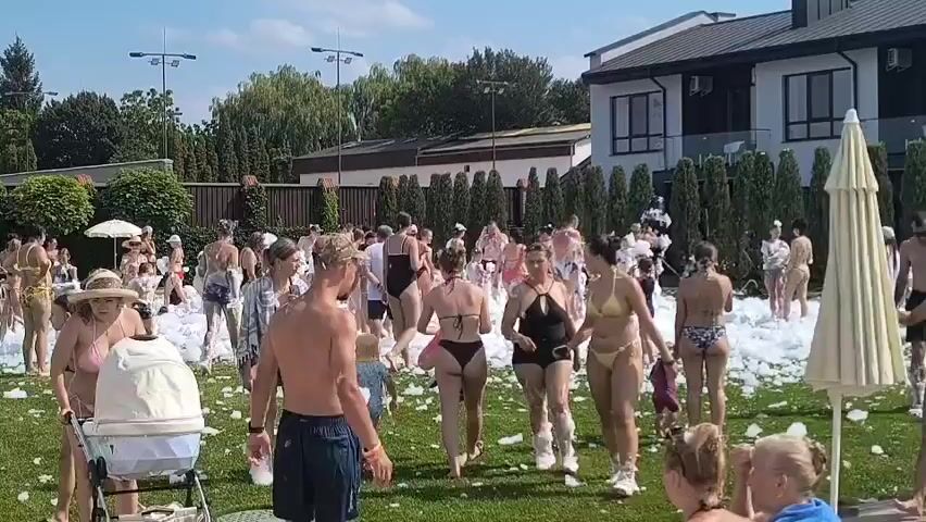 A large gathering of people, many in swimwear, are present outdoors on a grassy area. The ground and the people are covered in white foam, suggesting a foam party. There are several buildings in the background, as well as trees and a fence. Some people are walking, others are standing and interacting. One person is pushing a baby stroller. The sky is blue and appears to be a clear day.