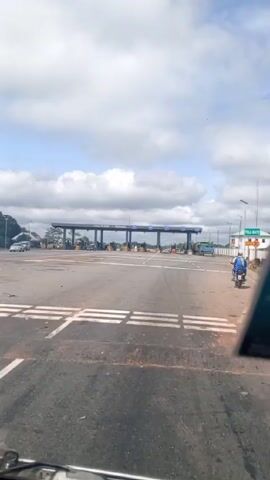 The view from inside a vehicle shows a paved highway approaching a toll plaza under a cloudy sky. A motorcyclist wearing blue clothes rides to the right side of the road, close to a green sign indicating “TOLL GATE 30”. The toll plaza has multiple lanes for vehicles to pass through.  Several vehicles are visible at the plaza. White lines are painted across the road in front of the camera.