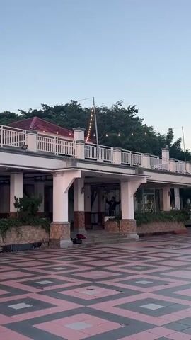 The video shows a wide shot of an outdoor area with a patterned tiled floor. In the background, there is a building with a white railing on the upper level and large pillars supporting it. Trees and a red roof are visible behind the building. Several people are present in the scene. One person is on a skateboard in the middle of the tiled area, appearing to perform a trick, while another person is seated on the tiled floor to the right. Two figures are near the entrance of the building, one of whom appears to be standing. The sky is clear and blue, indicating daytime.