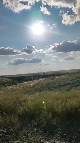 A wide shot captures a sun-drenched grassy field under a bright blue sky dotted with fluffy white clouds. In the distance, a bridge or overpass stretches across the landscape, with a few vehicles visible on it. The sun is high and bright, casting a lens flare in the center of the frame, and its rays illuminate the scene. The grass in the foreground appears dry and wispy, swaying slightly in what seems to be a gentle breeze. The overall impression is one of a warm, sunny day in a rural or open landscape.