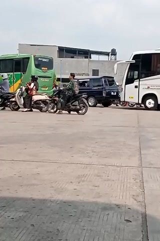 This media file shows an outdoor scene at what appears to be a gas station in Bandar Lampung, Indonesia. Several motorcycles and a large green bus are visible.

The primary subjects are people on motorcycles and the vehicles themselves. There are at least five motorcycles, with riders and passengers on some of them. The green bus has the text "ROYAL" and "Puspa Jaya" on its side, suggesting it's a passenger bus. A dark-colored SUV is parked behind the bus.

The activity depicted is people queuing or waiting at the gas station. Some individuals are seated on their motorcycles, while others appear to be interacting or waiting for service. The gas pumps are visible in the foreground, with a man standing near one of them.

Notable details include the bright, overcast sky, suggesting it is daytime. The concrete ground and the design of the gas station structures are typical of such facilities. The overall atmosphere appears functional and busy, with no strong emotions conveyed.

Visible text includes "ROYAL" and "Puspa Jaya" on the bus, and possibly "SOLAR" on one of the gas pumps, indicating diesel fuel. The scene is set outdoors, likely a public area.