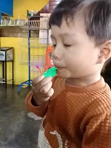 A young child with dark hair is holding a green and purple toy whistle in their mouth and blowing on it. The child is wearing an orange long-sleeved shirt with a dinosaur-like design on the front. The background appears to be indoors, with yellow and blue walls, shelving units with various items, and a refrigerator. The child's facial expression suggests concentration or effort.