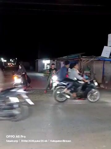 At night, on an outdoor road, there are several people on motorcycles facing the camera. Some of the people are wearing hijabs. A black SUV drives past on the right. Behind the motorcycles is a structure with a blue awning. The road is wet and shiny. A sign is visible on the right. Text on the screen indicates the video was recorded with an OPPO A6 Pro on May 31, 2026, in Kabupaten Bireuen.