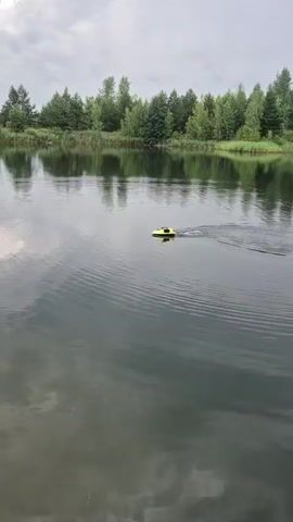 A remote-controlled boat with a yellow and black body is navigating on a calm body of water. The boat leaves a wake as it moves through the water, which is reflecting the surrounding trees and the overcast sky. The background consists of a line of lush green trees and vegetation along the edge of the water. The overall scene suggests an outdoor, natural setting, possibly a lake or pond, on a cloudy day.