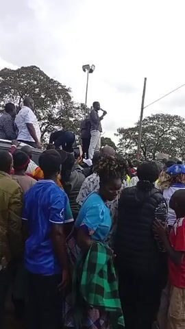 The video depicts a large crowd of people gathered outdoors. In the foreground, people are standing close together, some wearing casual clothing, others dressed in more traditional garments. Many are looking toward a group of individuals standing on the roof of a vehicle, likely a car or van. One person on the vehicle roof is holding a microphone and appears to be speaking to the crowd. The atmosphere seems lively. A large tree with broad branches is visible in the background, partially obscured by the crowd. A tall light pole is seen among the trees. The sky is overcast.