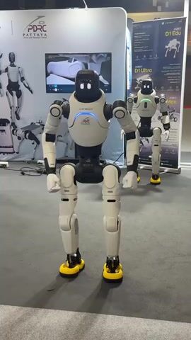 The video shows a humanoid robot with a white and black body, black head with glowing white eyes, and yellow feet. The robot is in a crouched position, with its arms extended forward and its hands open, appearing to be in a dance or demonstration. In the background, there are other robots, some standing and some in a similar crouched position. There are also displays with images of robots and text in what appears to be Thai. A person can be seen in the upper right corner, holding up a phone, possibly recording the robot. The environment appears to be an indoor exhibition or trade show.
