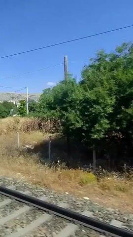 The video shows a view from a moving train. The train tracks are visible in the foreground. The landscape outside the train includes dry grass, bushes, and trees. There are also some white posts and what appear to be piles of building materials in the distance. A bridge crosses over the tracks. The sky is clear and blue. The camera appears to be moving horizontally, indicating the train is traveling along the tracks.