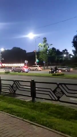 A nighttime, low-light video shows a street scene with cars and a motorcycle on a road. The sky is dark blue, and streetlights illuminate the area. In the background, a large sign with illuminated letters that appears to read "I <3 NOVOPOLOTSK" is visible. A black metal fence with a decorative pattern runs along the foreground, separating the sidewalk from the road. The sidewalk is paved with bricks. A few trees are present, and grass lines the area next to the road and sidewalk. A motorcycle with a rider is moving on the road, its taillight illuminated. Other vehicles are also present on the road, some appearing to be cars. The overall impression is of a city street at dusk or early night.