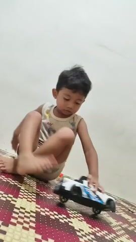 A young boy is playing with a toy car on a woven mat. He is wearing a light-colored tank top with a pattern and light-colored shorts. The boy is sitting on the mat with his legs bent and one foot extended towards the camera. He is holding the toy car with his right hand, and the car is white with blue and black accents. The background is a plain, light-colored wall. The video captures the boy's actions as he moves the toy car.