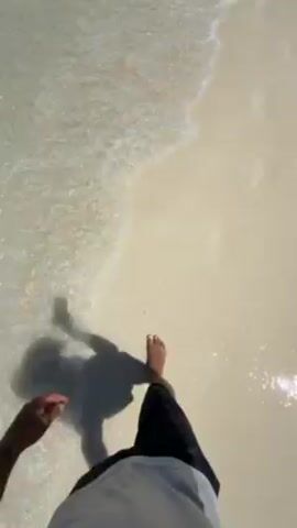 The video shows a first-person view of someone walking barefoot on a beach, with the clear, shallow water washing over the sand. The person is wearing dark shorts and a light-colored shirt. Their shadow is visible on the wet sand, and they are holding a phone in their hand as they walk. The sand is light in color, and the water is transparent.