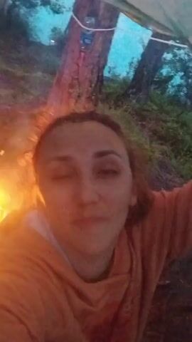 A selfie-style video shows a woman with curly hair and an orange hoodie. She winks and sticks out her tongue towards the camera. Behind her, a bonfire burns, illuminating the trees and foliage in the background. The lighting suggests it's dusk or nighttime. The woman appears to be outdoors, possibly camping or enjoying a fire in a natural setting.