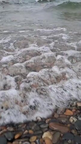 A video taken from a low angle shows a shoreline with a pebble beach and water. The water is clear enough to see the pebbles underneath. Small waves are rolling in and breaking on the shore, creating white foam. The sky is overcast and gray, and the water is a deep blue-gray color. In the distance, a hazy shoreline is visible, possibly with hills. The video appears to be shot on a cloudy day, creating a somber and tranquil atmosphere.
