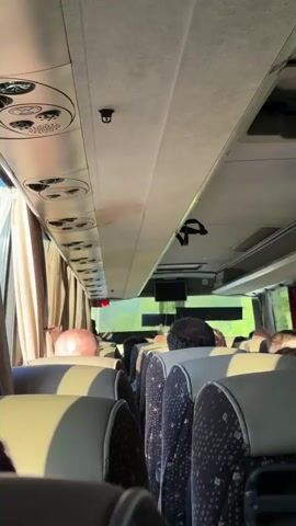 The video shows the interior of a bus from a passenger's perspective. The camera is pointed forward and slightly upward, capturing the ceiling of the bus and the backs of seats occupied by other passengers. The seats are upholstered in a patterned fabric with beige headrests. The ceiling is a light color, with air vents and lights visible. Curtains are drawn over the windows, revealing glimpses of green foliage outside. In the foreground, the backs of several headrests are visible. Further down the aisle, passengers' heads and shoulders are seen. One passenger in the middle of the bus raises their arm, reaching towards the ceiling. The lighting suggests it is daytime, possibly late afternoon due to the warm tones of the sunlight. The bus appears to be in motion, as indicated by the slightly blurred exterior scenery.
