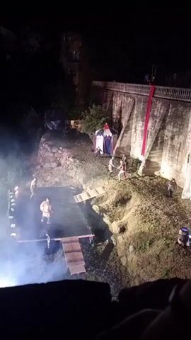 The video shows an outdoor nighttime performance. In the foreground, there is a wooden stage with a dark surface and a small wooden ramp leading to it. The stage is lit with bright white lights and filled with smoke. On the stage, there are two individuals dressed in what appear to be historical costumes, one in a more prominent position in the center. Several other people, also in costumes, are positioned on a slope to the right of the stage, near a tent and a large stone wall. The wall has a red banner hanging down its side. The overall impression is of a historical reenactment or theatrical performance taking place in a somewhat rugged, natural setting.